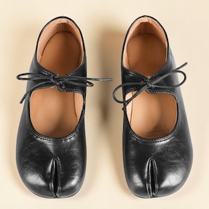 Op de afbeelding: Een paar zwarte leren Tabi-schoenen met een split-teenontwerp. De schoenen hebben een Mary Jane-stijl met een bandje en striksluiting. De binnenkant is lichtbruin.