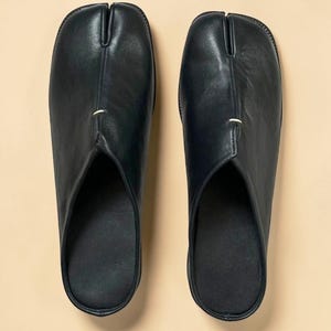 May include: A pair of black leather split-toe slippers. The slippers have a distinctive design with a separation for the big toe. The footwear is a slip-on style, with a smooth, matte finish. The slippers are shown on a neutral background.