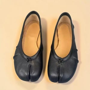 Puede incluir: Un par de bailarinas negras con puntera dividida. Los zapatos tienen una puntera redondeada con una divisi&oacute;n para el dedo gordo, un peque&ntilde;o detalle de lazo y una plantilla beige. Las bailarinas est&aacute;n hechas de un material liso y mate.