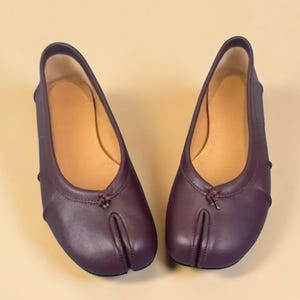 Puede incluir: Un par de bailarinas de cuero color ciruela con dise&ntilde;o de puntera dividida. Los zapatos tienen una puntera redondeada y un peque&ntilde;o lazo decorativo en la parte superior. El interior es de color beige claro.