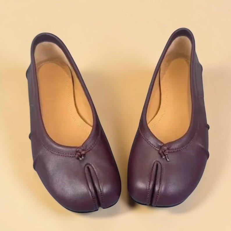 Puede incluir: Un par de bailarinas de cuero color ciruela con dise&ntilde;o de puntera dividida. Los zapatos tienen una puntera redondeada y un peque&ntilde;o lazo decorativo en la parte superior. El interior es de color beige claro.