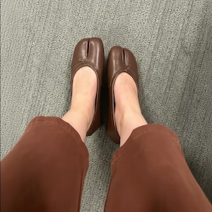 Puede incluir: Bailarinas marrones de cuero con puntera dividida. Los zapatos tienen una puntera redondeada con un peque&ntilde;o lazo. Las bailarinas se llevan con pantalones marrones. Zapatos de estilo &uacute;nico.