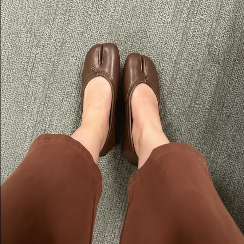 Puede incluir: Bailarinas marrones de cuero con puntera dividida. Los zapatos tienen una puntera redondeada con un peque&ntilde;o lazo. Las bailarinas se llevan con pantalones marrones. Zapatos de estilo &uacute;nico.