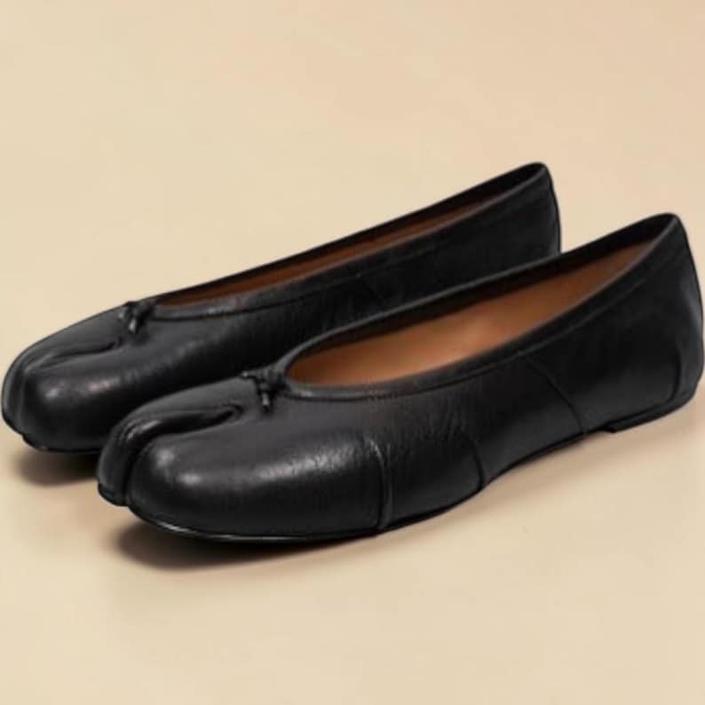 Puede incluir: Un par de bailarinas de cuero negro. Los zapatos tienen un dise&ntilde;o de punta dividida y un peque&ntilde;o lazo en la parte superior. Las bailarinas est&aacute;n sobre un fondo neutro.