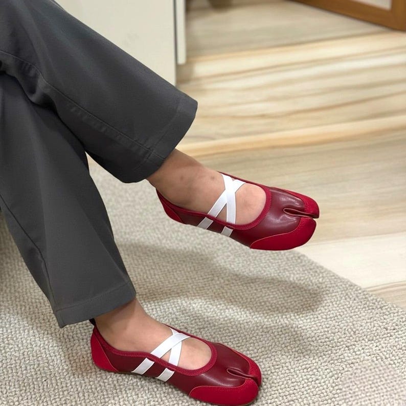 May include: Red split-toe shoes with white crisscross straps. The shoes have a rounded toe and a contrasting red suede-like material on the toe and heel. The shoes are designed to be worn with the big toe separated from the other toes.