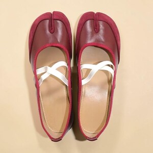 May include: A pair of burgundy split-toe ballet flats with white criss-cross straps. The shoes have a leather upper with a suede toe cap and a red trim. The soles are a light tan color.