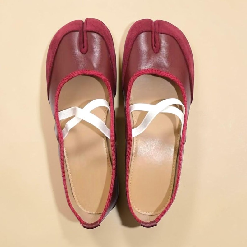 May include: A pair of burgundy split-toe ballet flats with white criss-cross straps. The shoes have a leather upper with a suede toe cap and a red trim. The soles are a light tan color.