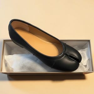 Puede incluir: Bailarinas de cuero negro con dise&ntilde;o de punta dividida, presentadas en una caja. Los zapatos tienen una textura suave y una plantilla marr&oacute;n claro. La caja es beige y contiene papel de seda blanco.