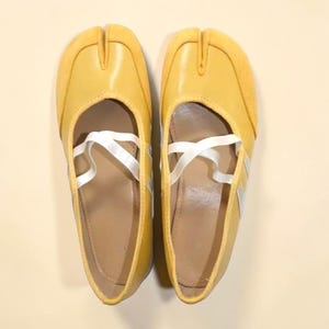 May include: A pair of yellow split-toe ballet flats with white crisscross straps. The shoes have a rounded toe and a small heel. The upper is a solid yellow, and the sole is a light brown color. The shoes are designed to be worn by dancers.