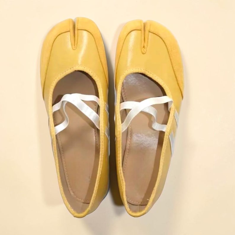 May include: A pair of yellow split-toe ballet flats with white crisscross straps. The shoes have a rounded toe and a small heel. The upper is a solid yellow, and the sole is a light brown color. The shoes are designed to be worn by dancers.