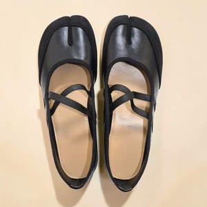 May include: A pair of black split-toe shoes with a strap design. The shoes have a rounded toe and a strap that crosses over the top of the foot. The shoes are made of leather and have a suede-like material on the toe.