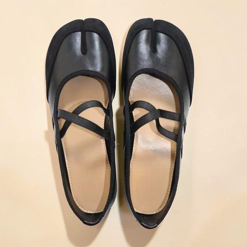 May include: A pair of black split-toe shoes with a strap design. The shoes have a rounded toe and a strap that crosses over the top of the foot. The shoes are made of leather and have a suede-like material on the toe.