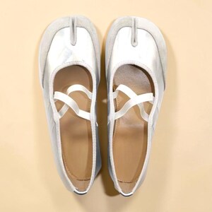 May include: A pair of silver ballet flats with a split toe design, featuring a light gray suede trim and white crisscross straps. The shoes have a light brown insole and a rounded toe.