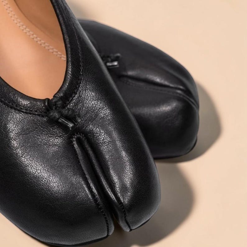 Puede incluir: Bailarinas de cuero negro con dise&ntilde;o de puntera dividida. Los zapatos tienen una puntera redondeada y un peque&ntilde;o detalle de lazo en la parte superior. Las bailarinas son de cuero liso y texturizado.