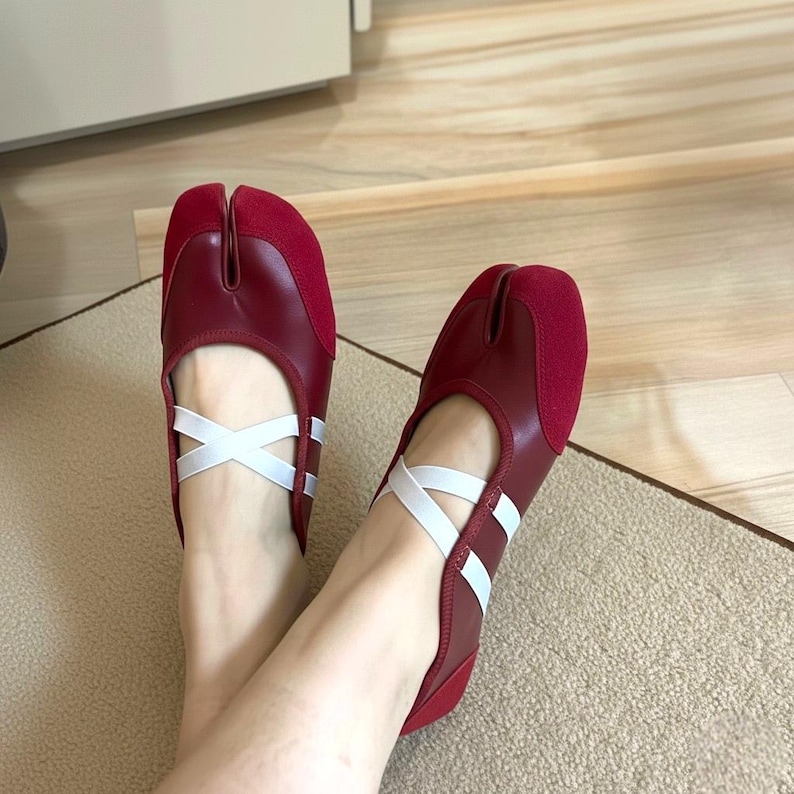 May include: A pair of burgundy Tabi shoes with white criss-cross straps. The shoes have a split toe design and a suede-like material on the top of the foot. The soles are a darker shade of red.