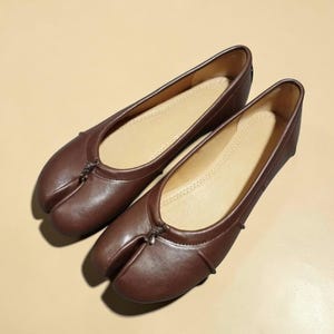 Puede incluir: Un par de bailarinas de cuero marr&oacute;n con dise&ntilde;o de puntera dividida. Los zapatos tienen una puntera redondeada y un peque&ntilde;o nudo decorativo en la parte superior. El interior es de color beige claro.