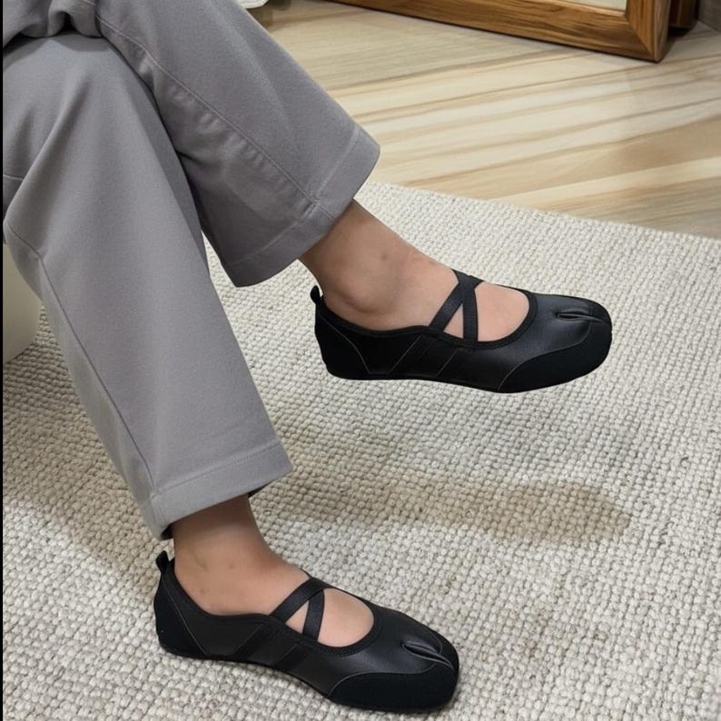 May include: Black split-toe ballet flats with crisscross straps. The shoes have a low heel and a unique design that separates the big toe. The flats are made of a smooth, matte material.