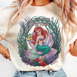 May include: Cream-colored t-shirt featuring a colorful graphic of Ariel from The Little Mermaid. The design includes Ariel, Flounder, Sebastian, and the words "The Little Mermaid" in a decorative font, surrounded by aquatic plants.