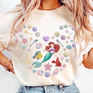 May include: Ivory-colored t-shirt featuring a mermaid design surrounded by seashells, starfish, and other sea-themed illustrations. The mermaid has red hair and a teal tail. The shirt also has the word "Colors" and "Ivory" printed on it.