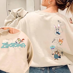 May include: A beige crewneck sweatshirt featuring Zootopia characters. The front displays the Zootopia logo with Judy Hopps and Nick Wilde. The back shows a vertical design with Judy, Nick, Flash, and Mr. Big connected by a dotted line with icons.