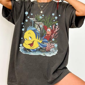Puede incluir: Camiseta gris oscuro con una ilustraci&oacute;n de dibujos animados de Flounder y Sebasti&aacute;n de La Sirenita. El dise&ntilde;o incluye burbujas azules, coral y algas marinas. La camiseta tiene mangas cortas y un corte holgado.