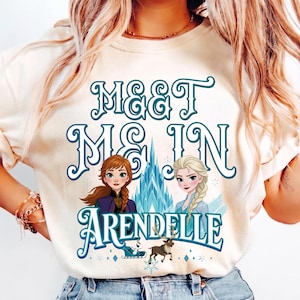 May include: Cream-colored t-shirt with the text "MEET ME IN ARENDELLE" in teal script. The graphic features illustrations of two princesses, a castle, and a reindeer. The shirt is made of a soft material.