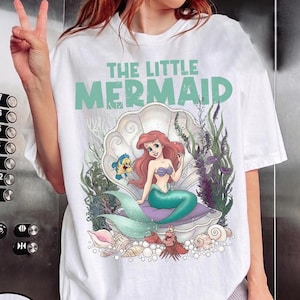 May include: White t-shirt featuring a graphic of Ariel from The Little Mermaid. The design includes Ariel sitting in a seashell, with Flounder and Sebastian. The text "THE LITTLE MERMAID" is printed above the image in a teal color.