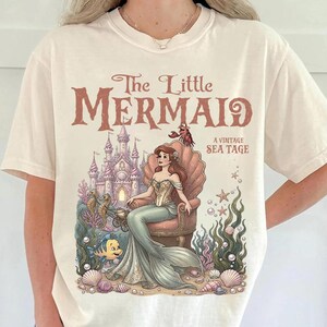 May include: Cream-colored t-shirt featuring a vintage-style illustration from "The Little Mermaid." The design includes the title, a mermaid seated on a shell throne, a castle, and sea creatures. The shirt has short sleeves and a crew neck.
