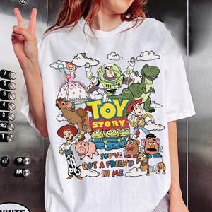 Op de afbeelding: Wit T-shirt met een kleurrijke afbeelding van personages uit de film "Toy Story". Het ontwerp bevat Buzz Lightyear, Woody en andere personages, met de tekst "TOY STORY" en "YOU'VE GOT A FRIEND IN ME."