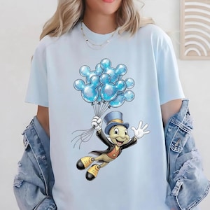 May include: Light blue t-shirt featuring a cartoon character holding a bunch of blue Mickey Mouse-shaped balloons. The character is wearing a top hat, black jacket, and yellow shoes. The balloons have a glittery effect.
