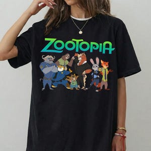 K&ouml;nnte beinhalten: Schwarzes T-Shirt mit dem Wort "ZOOTOPIA" in gr&uuml;nen und blauen Farbverlauf-Buchstaben. Das Shirt zeigt Zeichentrickfiguren aus dem Film, darunter einen Polizisten, ein Faultier und einen Fuchs.