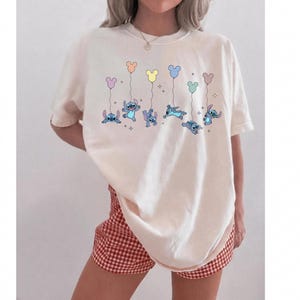 May include: An off-white short-sleeve t-shirt featuring a graphic print of the blue character Stitch in various poses, each holding a pastel-colored balloon shaped like a mouse head, accompanied by small star accents scattered across the chest area.