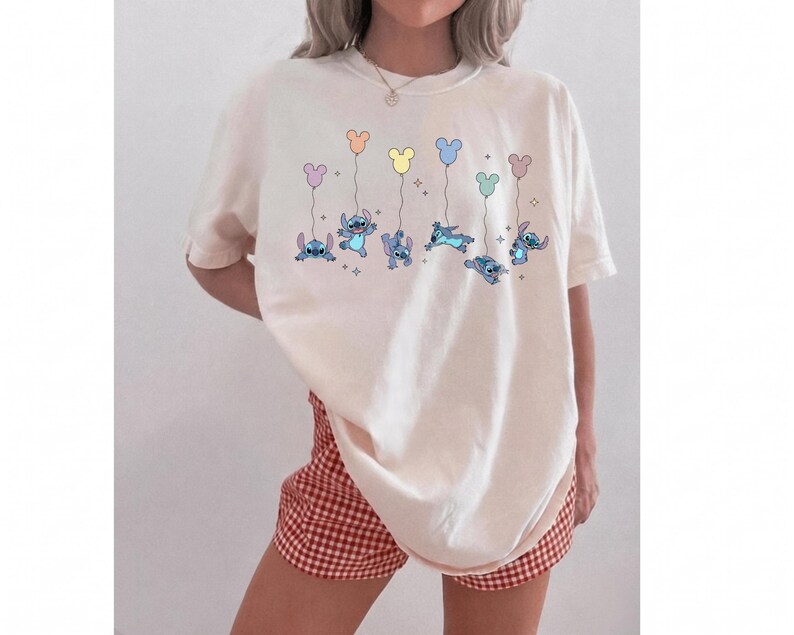 May include: An off-white short-sleeve t-shirt featuring a graphic print of the blue character Stitch in various poses, each holding a pastel-colored balloon shaped like a mouse head, accompanied by small star accents scattered across the chest area.