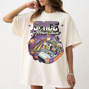 May include: Cream-coloured t-shirt with a vibrant graphic of Mickey Mouse, Donald Duck, Goofy, and Pluto on a roller coaster. The design includes the words "SPACE MOUNTAIN" in purple and white, with stars and planets.