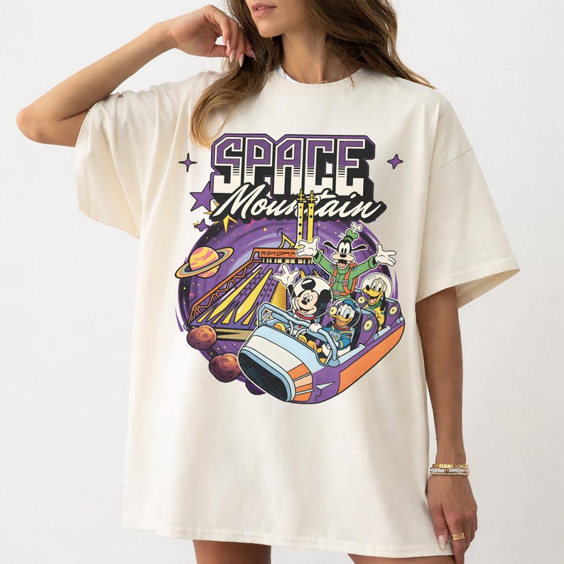 May include: Cream-coloured t-shirt with a vibrant graphic of Mickey Mouse, Donald Duck, Goofy, and Pluto on a roller coaster. The design includes the words "SPACE MOUNTAIN" in purple and white, with stars and planets.