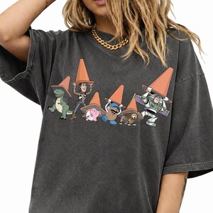 May include: Dark gray t-shirt featuring a graphic of Toy Story characters wearing orange traffic cones. The design includes Rex, Woody, Hamm, Mr. Potato Head, Slinky Dog, and Buzz Lightyear. The shirt has short sleeves and a relaxed fit.
