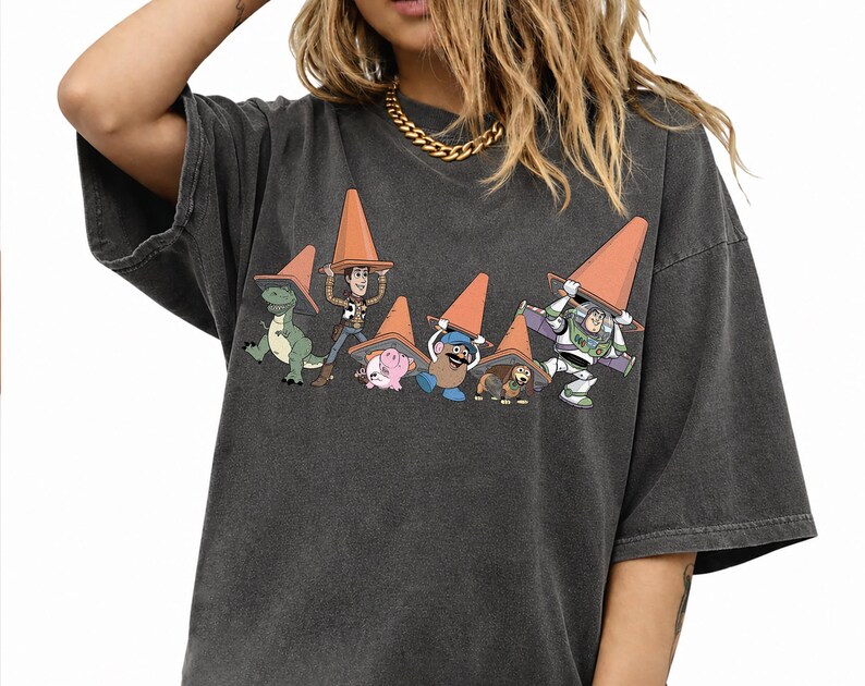 May include: Dark gray t-shirt featuring a graphic of Toy Story characters wearing orange traffic cones. The design includes Rex, Woody, Hamm, Mr. Potato Head, Slinky Dog, and Buzz Lightyear. The shirt has short sleeves and a relaxed fit.