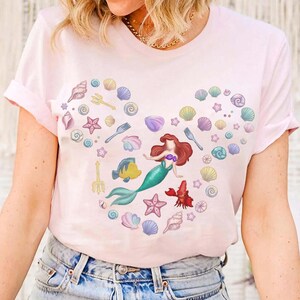 May include: Light pink t-shirt featuring a heart-shaped design composed of seashells, starfish, and a mermaid illustration. The mermaid has red hair and a teal tail. The design includes a yellow fish and a red crab. The shirt is made of a soft material.