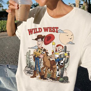 Pu&ograve; includere: T-shirt color crema con un design del West selvaggio con personaggi dei cartoni animati. La grafica include Woody, Jessie, un cavallo e uno scenario desertico. Il testo "WILD WEST" &egrave; stampato in rosso sopra i personaggi.