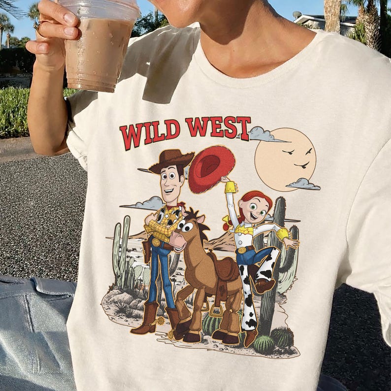Pu&ograve; includere: T-shirt color crema con un design del West selvaggio con personaggi dei cartoni animati. La grafica include Woody, Jessie, un cavallo e uno scenario desertico. Il testo "WILD WEST" &egrave; stampato in rosso sopra i personaggi.