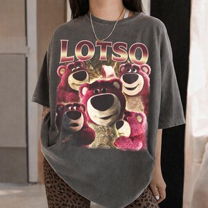 May include: Dark gray t-shirt featuring a graphic of Lotso, the bear from Toy Story, with the word "LOTSO" in a gold and red font. The image shows Lotso and his friends. The shirt has short sleeves.