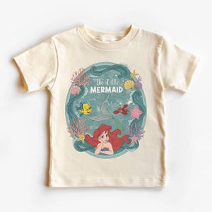 May include: A cream-colored t-shirt featuring a circular graphic of Ariel from The Little Mermaid. The graphic includes Ariel, Flounder, Sebastian, and the text "The Little Mermaid" surrounded by coral and seashells.