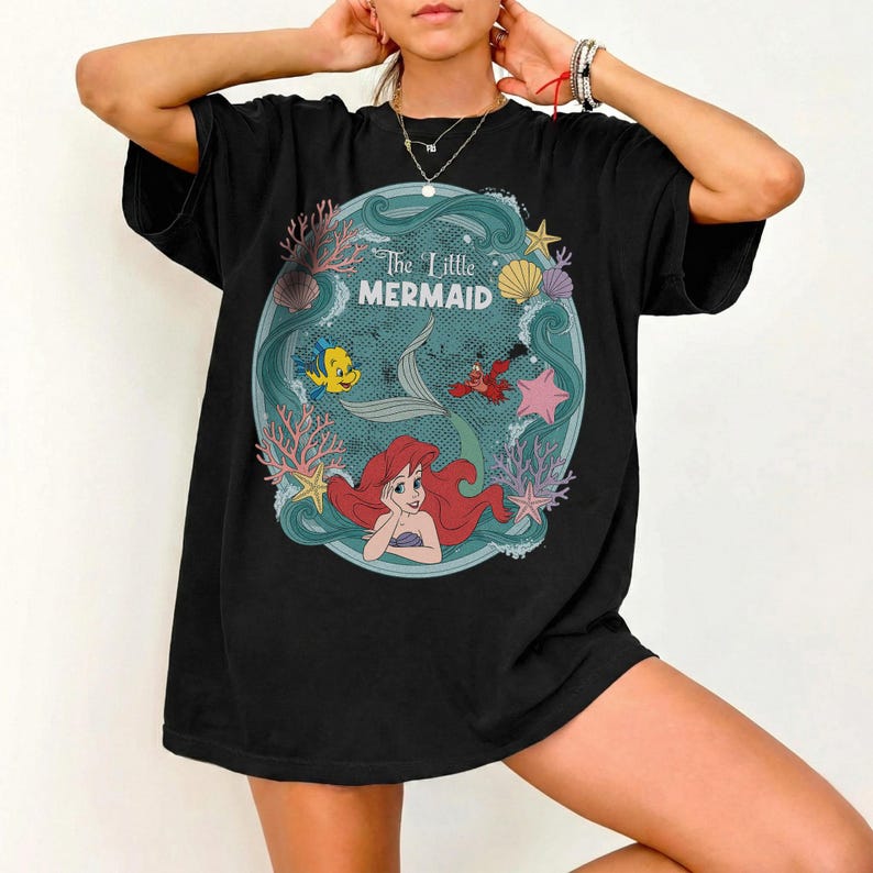 May include: Black t-shirt featuring a circular design with the text "The Little Mermaid." The design includes Ariel, Flounder, Sebastian, and other sea creatures. The shirt has short sleeves and a relaxed fit.