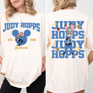 May include: An off-white t-shirt featuring Judy Hopps from Zootopia. The front displays the text Judy Hopps, Est. 2016, Zootopia, and a graphic of the character inside a blue Mickey Mouse icon. The back shows Judy Hopps with a badge and carrot.
