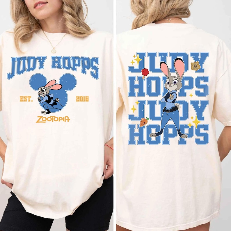 May include: An off-white t-shirt featuring Judy Hopps from Zootopia. The front displays the text Judy Hopps, Est. 2016, Zootopia, and a graphic of the character inside a blue Mickey Mouse icon. The back shows Judy Hopps with a badge and carrot.