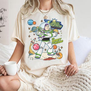 May include: Cream-colored t-shirt featuring a colorful graphic of Buzz Lightyear from Toy Story. The design includes the text "Buzz Lightyear Laser Blast" and "To Infinity and Beyond" with planets, stars, and rockets.
