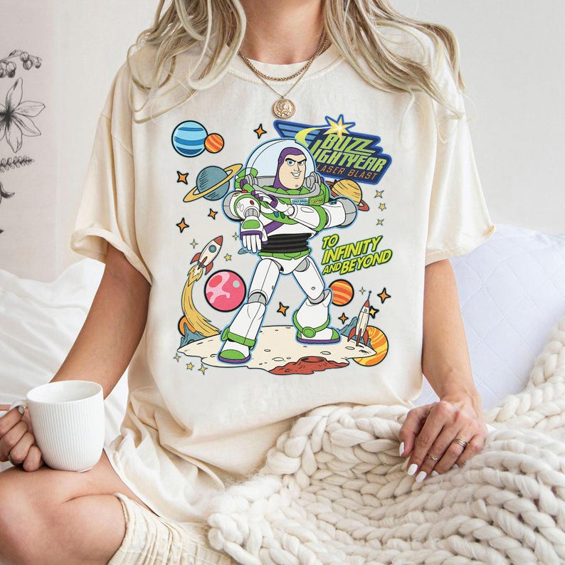 May include: Cream-colored t-shirt featuring a colorful graphic of Buzz Lightyear from Toy Story. The design includes the text "Buzz Lightyear Laser Blast" and "To Infinity and Beyond" with planets, stars, and rockets.