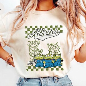 May include: Ivory-colored t-shirt featuring a graphic of green aliens in blue overalls, with the word "Aliens" above them and the year "1995" below. The design is set against a green and white checkered background.