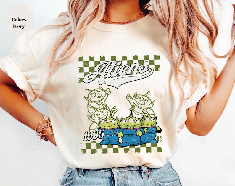 May include: Ivory-colored t-shirt featuring a graphic of green aliens in blue overalls, with the word "Aliens" above them and the year "1995" below. The design is set against a green and white checkered background.