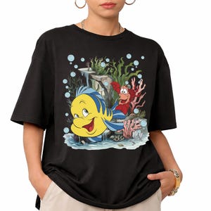 Puede incluir: Camiseta negra con un gr&aacute;fico colorido de Flounder, Sebasti&aacute;n y un paisaje submarino de La Sirenita. El dise&ntilde;o incluye burbujas y coral. La camiseta tiene un ajuste casual y holgado.